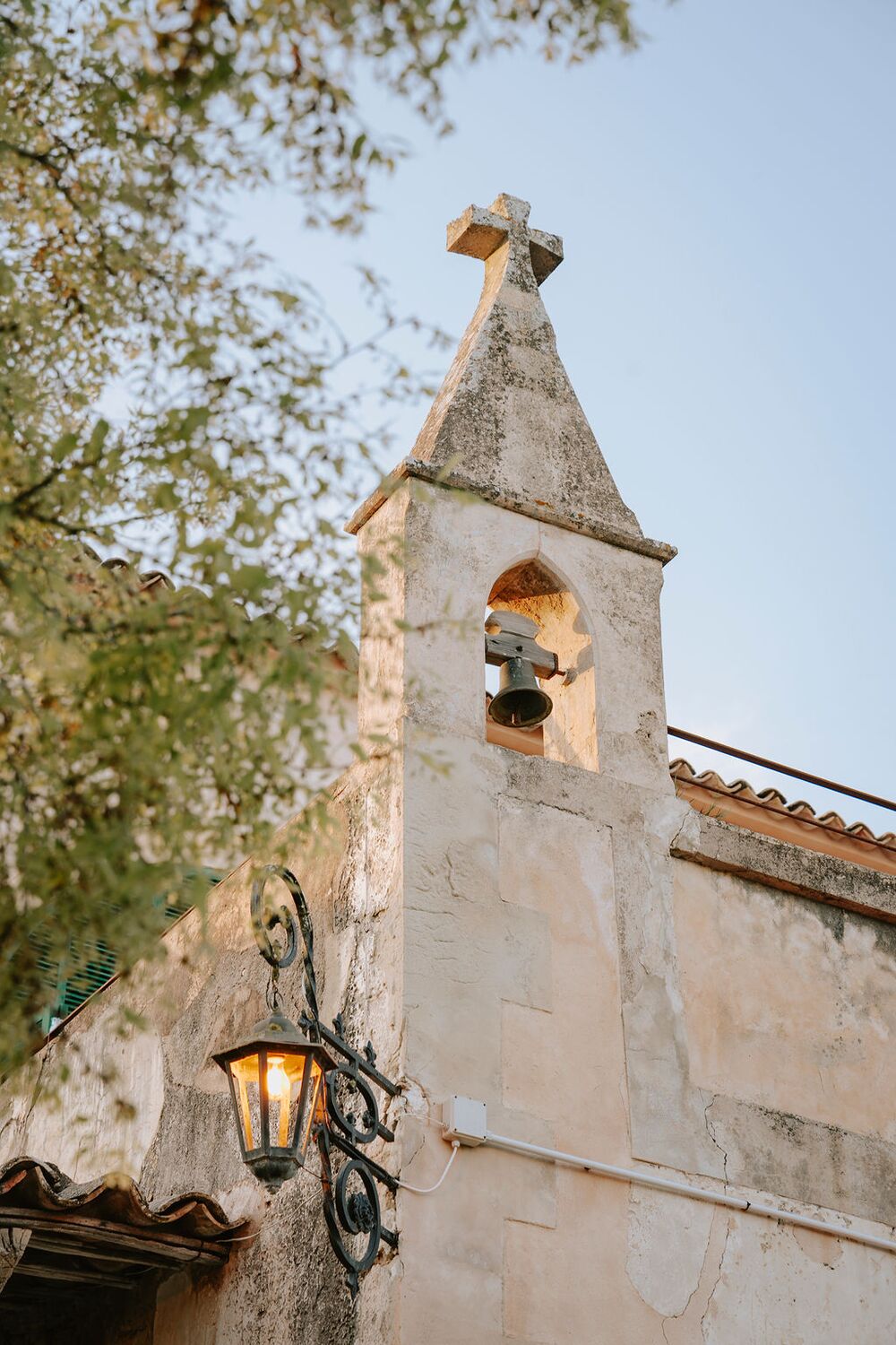 The stone bell-gable of the manor crowned with a cross, an old wrought-iron lantern below it against the evening sky