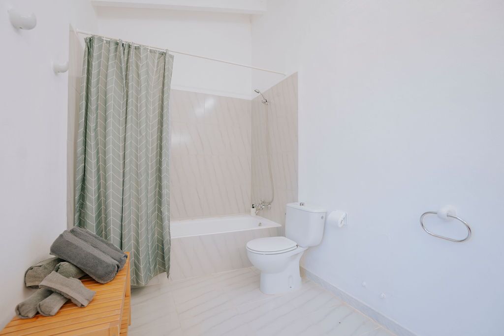 A bright white bathroom with a green-patterned shower curtain and folded towels on a wooden bench